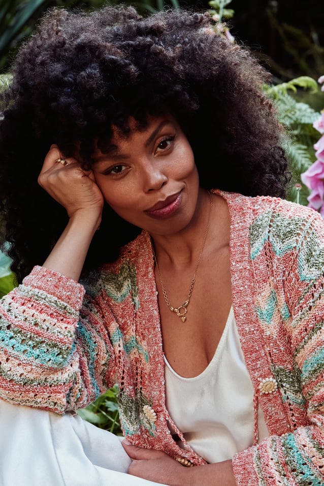 Emma Dabiri in white top with pastel coloured knit cardigan sat in a forest amongst pink foxglove flowers. She is wearing statement earrings, necklace and Claddagh ring.