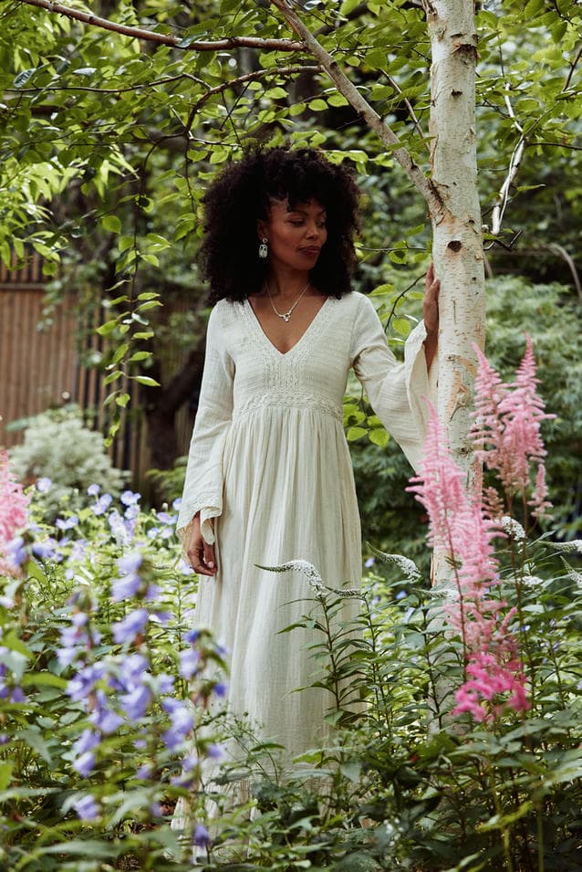 Emma Dabiri in white dress next to a silver birch in the forest, wearing silver statement earrings and necklace.