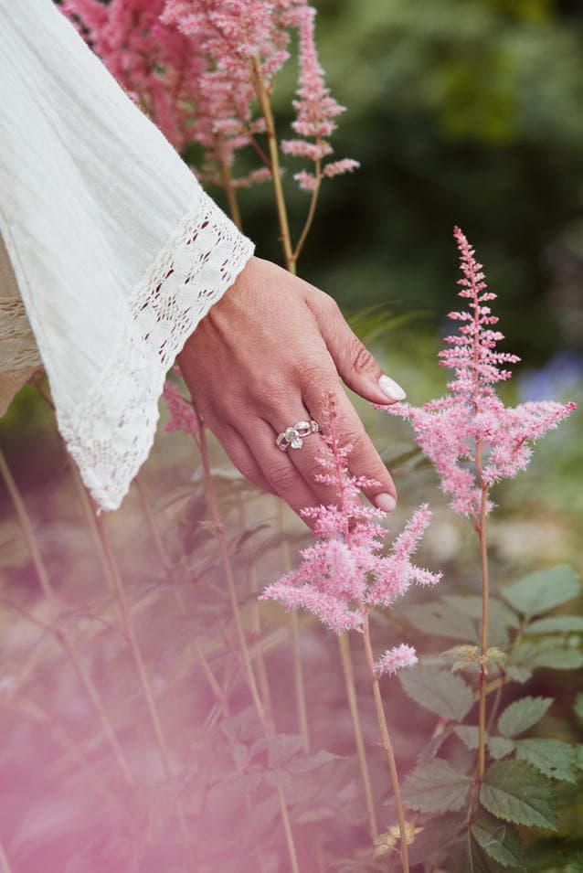 Womans hand gently swaing pink flowers, wearing white flowy long sleaved dress and silver claddagh ring